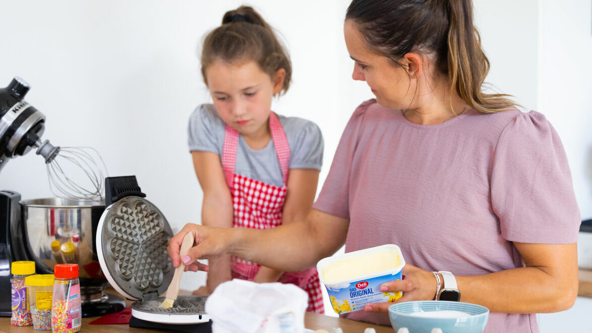Eine Frau fettet ein Waffeleisen mit Margarine beim gemeinsamen Backen mit ihrer Tochter.